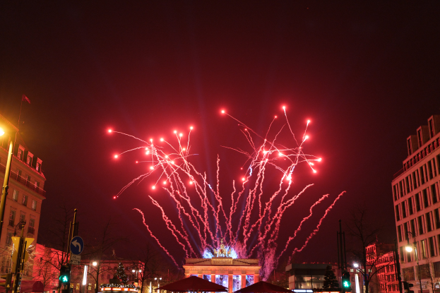 Eine Straße in Berlin an Silvester, gefüllt mit Gebäuden, Bäumen, Laternenmasten, Verkehrsschildern, Zelten und Menschen, mit einem von Feuerwerk erleuchteten Himmel im Hintergrund.