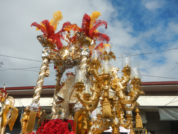 Ein großes goldenes und rotes Festwagen, geschmückt mit Blumen und anderen Dekorationen bei einem Faschingsumzug, mit einem Gebäude, Strommasten mit Drähten und einem bewölkten Himmel im Hintergrund.