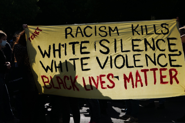 Eine Gruppe maskierter Menschen hält ein Banner mit der Aufschrift "Rassismus tötet Weiße Stille, weiße Gewalt, Schwarze Leben zählen" auf einer dunklen Straße.