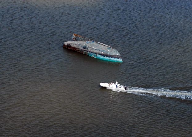 Ein kleines, teilweise unter Wasser liegendes Wrackboot, das neben einem größeren Boot auf einem Fluss fährt, mit ein paar Menschen an Bord.