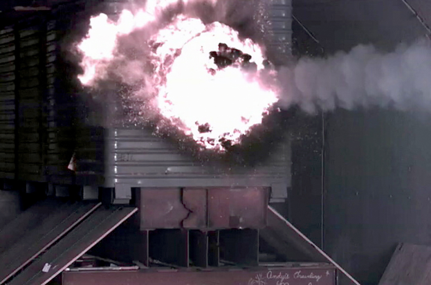 Eine große Explosion in der Mitte eines Gebäudes mit Rauch, der herausquillt, eine Wand im Hintergrund und metallische Gegenstände mit einer Tafel unten.