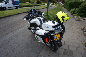 Ein auf der Straße geparktes Polizeimotorrad mit einem Helm darauf, umgeben von Bäumen, Pfählen, Gebäuden, Pflanzen, Gras, Steinen und anderen Fahrzeugen.