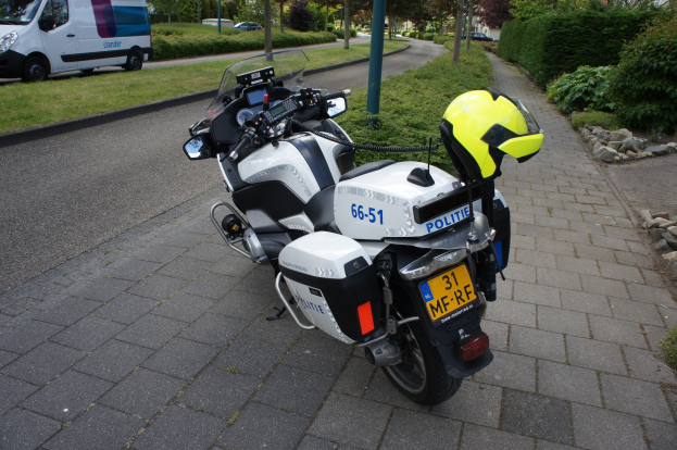 Ein auf der Straße geparktes Polizeimotorrad mit einem Helm darauf, umgeben von Bäumen, Pfählen, Gebäuden, Pflanzen, Gras, Steinen und anderen Fahrzeugen.