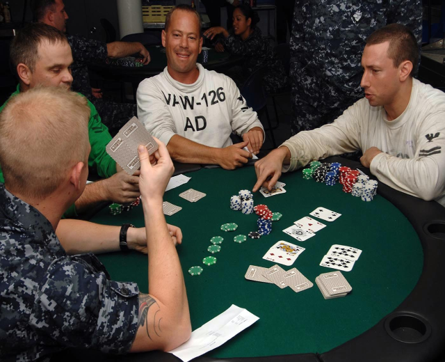 Eine Gruppe von Männern, die Poker spielen, an einem Tisch mit Münzen und Karten vor sich, in einer turnierähnlichen Umgebung mit einer Säule im Hintergrund.