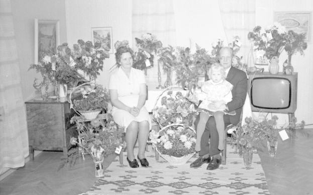 Schwarzes und weißes Foto einer Familie aus drei Personen - einem Mann, einer Frau und einem Kind - die auf einem Sofa vor einem Fernseher sitzen, Blumenvasen auf den Seitentischen, einem Teppich auf dem Boden, Vorhängen im Hintergrund und Fotorahmen an der Wand.