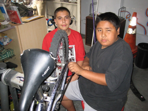 Zwei Personen reparieren ein Fahrrad in der Mitte, während Büromaterialien und Fahrräder im Hintergrund sichtbar sind.