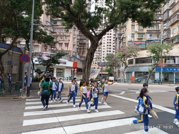 Eine Gruppe von Kindern in blauen und weißen Uniformen überquert eine Straße, die von Bäumen, Laternenmasten und Schildern gesäumt ist, mit hohen Gebäuden und einem sichtbaren Himmel im Hintergrund.