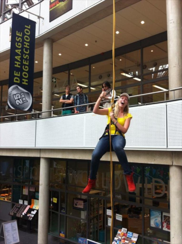 Eine Frau in einem gelben T-Shirt, einer Mütze und roten Stiefeln hängt an einem Seil in einem Raum mit Bücherregalen, einer Tafel, einem Banner, mehreren Personen, Glastüren und Deckenleuchten.