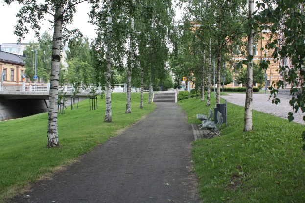 Ein parkartiger Weg mit Bäumen und Bänken, Grünfläche, Brücke mit Geländer, Gebäuden im Hintergrund, Treppen, Laternen und Himmel.