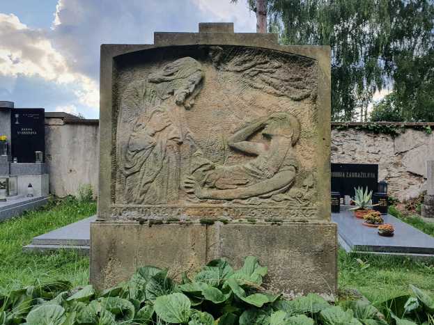 Ein steinernes Denkmal mit einer Skulptur einer Frau und eines Kindes, umgeben von Pflanzen am Boden, einer Wand im Hintergrund und Bäumen und Wolken am Himmel.