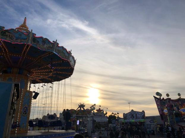 Ein Karussell auf einem Rummelplatz mit Menschen darauf, umgeben von Fahrgeschäften, Pfählen und Lichtern, unter einem bewölkten Himmel, der von der untergehenden Sonne erhellt wird.