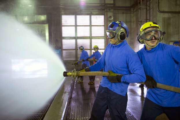 Eine Gruppe von Männern in blauen Hemden und gelben Helmen arbeitet an einer Maschine in einer Fabrikumgebung, wobei einer der Männer den Boden mit einem Schlauch bespritzt.