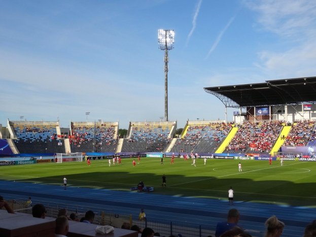 Ein Fußballspiel in einem großen Stadion mit Zuschauern auf den Rängen und Spielern auf dem Feld, unter Flutlicht mit dem Himmel im Hintergrund.
