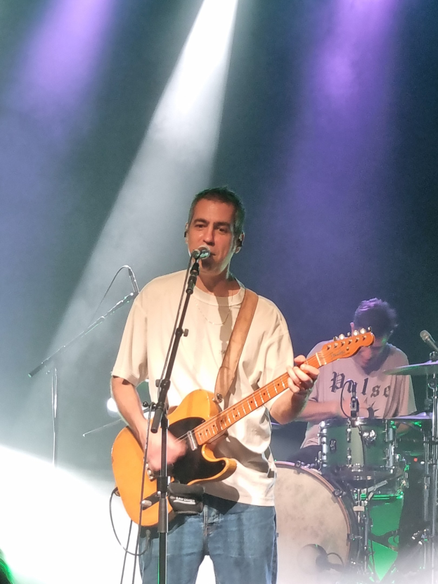 Ein Mann spielt eine akustische Gitarre und singt in ein Mikrofon auf der Bühne, während ein anderer Mann Schlagzeug spielt.