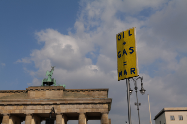 Das Brandenburgische Tor in Berlin, Deutschland, mit einem gelben Schild, das "Öl- und Gaskrieg" lautet, Gebäuden, Polen, Lampen, einer Statue und einem bewölkten Himmel im Hintergrund.