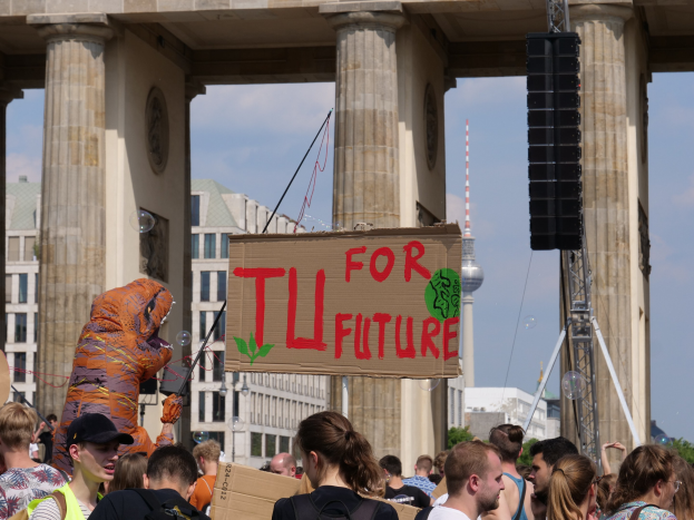 Eine Gruppe von Menschen steht vor dem Brandenburger Tor in Berlin, Deutschland, wobei einige Taschen tragen und eine Person in einem Kostüm mit einer Tafel steht, vor einem Hintergrund von Gebäuden, Bäumen und einem Turm unter einem bewölkten Himmel.