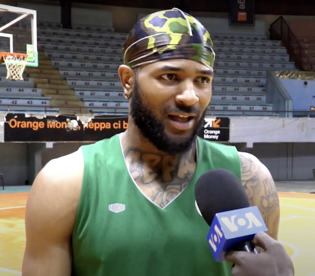 Ein Mann in einem grünen Tanktop, der in ein Mikrofon spricht, vor einem Basketballfeld mit einem Netz und Treppen im Hintergrund.