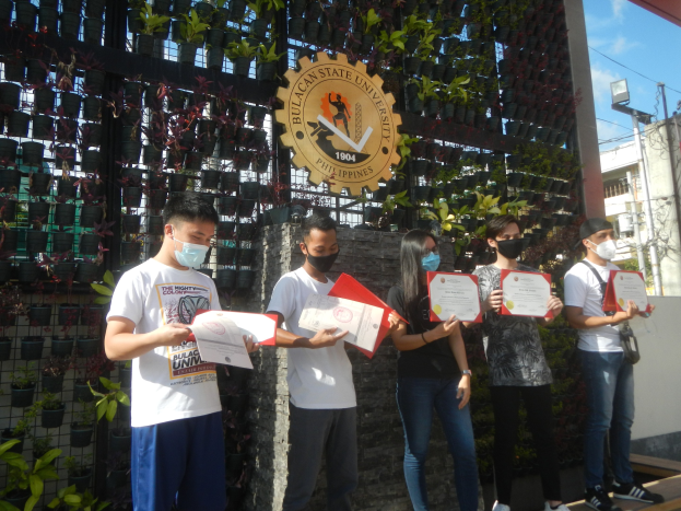 Gruppe von maskierten Menschen mit Urkunden vor einem Gebäude mit bepflanzter Wand links und einem gebogenen Logo rechts, mit städtischem Hintergrund.