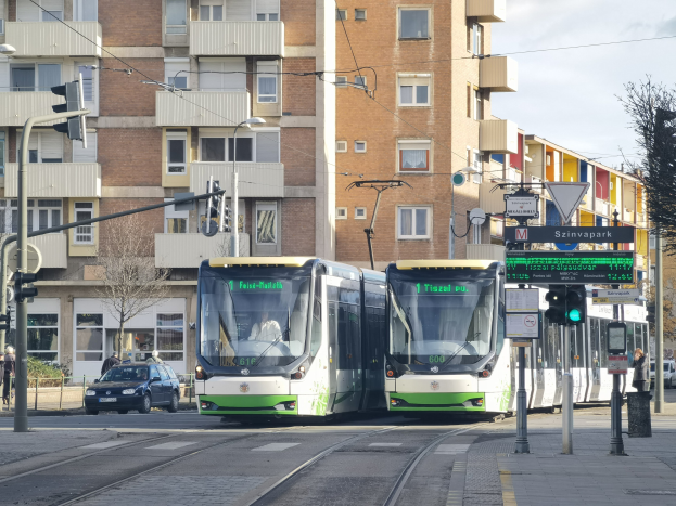 Zwei grüne und weiße Straßenbahnen auf einer Stadtstraße mit hohen Gebäuden, Fahrzeugen, Fußgängern, Verkehrszeichen und Bäumen unter einem bewölkten Himmel.