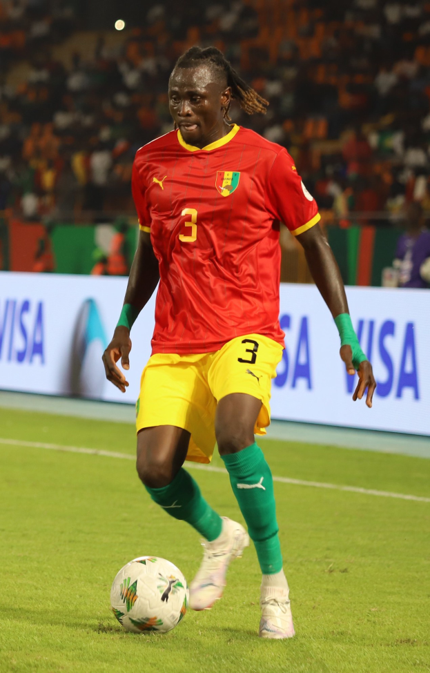 Ein Mann in roter und gelber Uniform spielt Fußball auf einem Feld, umgeben von einer Zuschauermenge auf den Tribünen und Werbetafeln im Hintergrund.