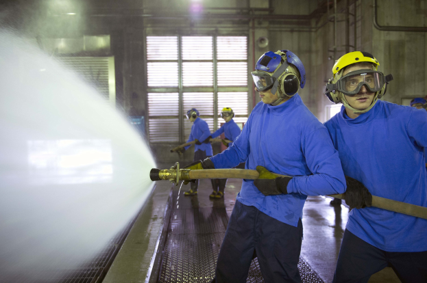 Eine Gruppe von Männern in blauen Hemden und gelben Helmen arbeitet an Maschinen, wobei einer einen Wasserstrahl auf den Boden sprüht, in einer Fabrikumgebung mit sichtbaren Rohren, Fenstern und Lichtern.