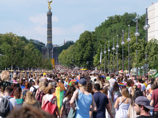 Eine große Menschenmenge geht eine Straße in Berlin, Deutschland, entlang, mit Laternen an der Straße, Bäumen und einem Turm mit einer Statue im Hintergrund und Gebäuden unter einem bewölkten Himmel.