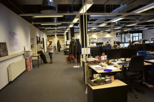 Ein großer Open-Plan-Büroraum mit Menschen, die an Schreibtischen arbeiten, Computern und Monitoren, umgeben von Stühlen, Papieren, Büchern und Kartons, mit Kleidung in der Nähe und Informationsplakaten an den Wänden unter Deckenleuchten und Glasfenstern.