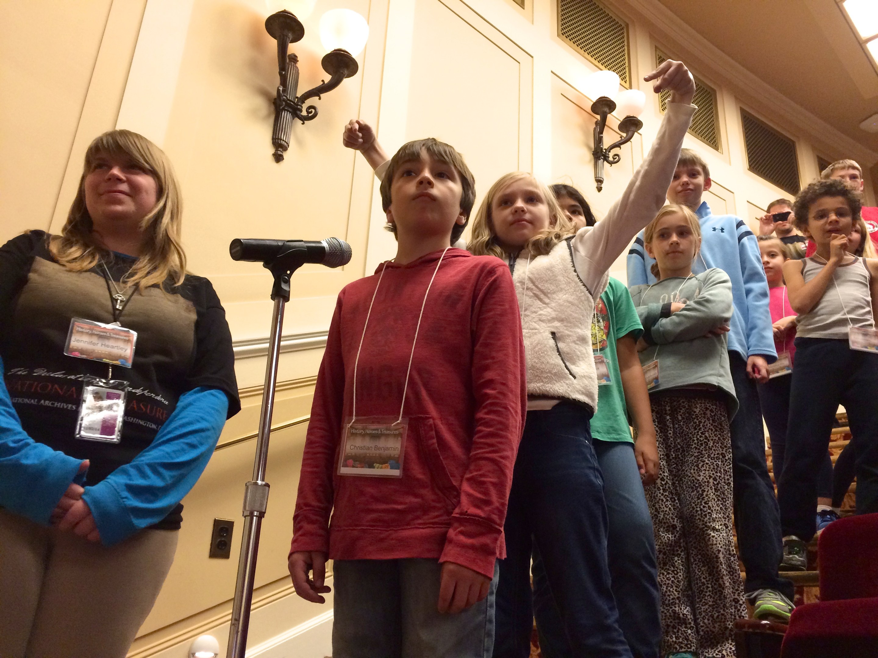 Gruppe von Kindern, die an einem Mikrofon singen, mit einer Frau neben ihnen, Stühlen auf der rechten Seite und Wandlampen im Hintergrund.