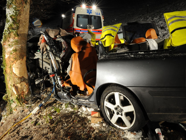 Auto auf der Seite einer schneebedeckten Straße mit einem Krankenwagen im Hintergrund, umgeben von Bäumen.