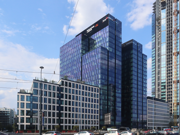 Ein großes Gebäude mit vielen Fenstern, das als Hauptsitz der Deutschen Bank identifiziert wurde, steht in der Mitte einer Stadt umgeben von Straßenmasten, Straßenlaternen, Strommasten, Stromkabeln, Kraftfahrzeugen, Büschen, Bäumen und einem bewölkten Himmel.