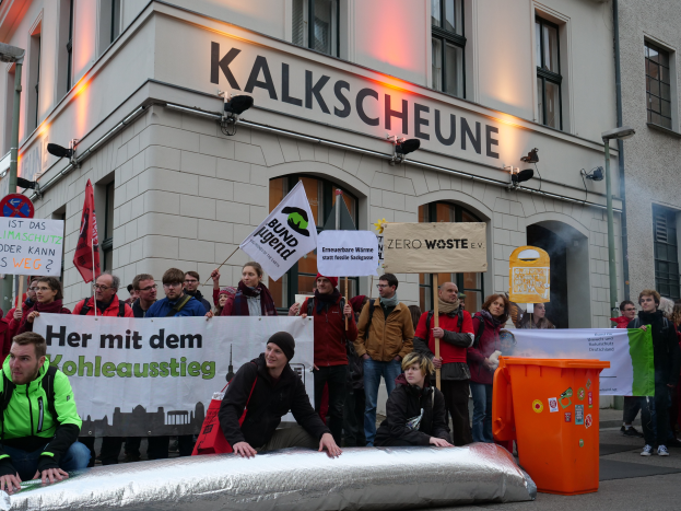 Eine Gruppe von Menschen mit Schildern und Plakaten steht vor einem Gebäude, mit zwei Personen im Vordergrund und einem Müllcontainer auf der rechten Seite während einer Demonstration in Deutschland.