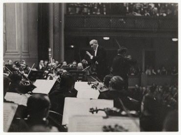 Schwarzes und weißes Foto eines Dirigenten vor einem Orchester, der einen Taktstock hält, mit sitzenden Musikern, die Instrumente spielen und Notenständer mit Büchern im Vordergrund.