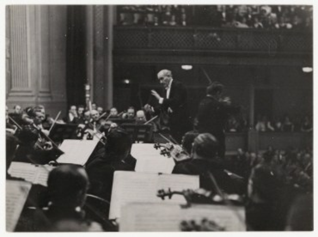Schwarzes und weißes Foto eines Dirigenten vor einem Orchester, der einen Taktstock hält, mit sitzenden Musikern, die Instrumente spielen und Notenständer mit Büchern im Vordergrund.