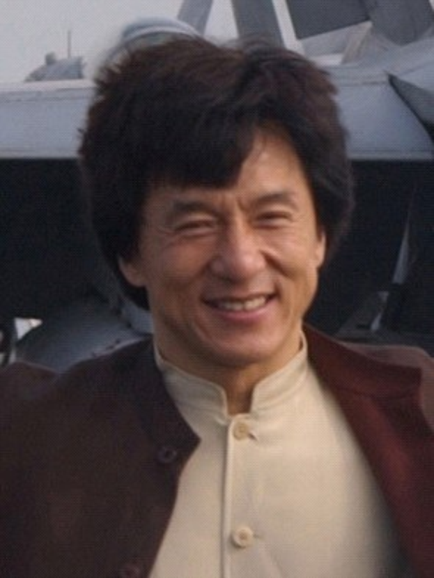 Jackie Chan steht strahlend vor einem Flugzeug unter einem klaren blauen Himmel, trägt ein weißes Hemd und eine schwarze Jacke mit ordentlich gekämmtem Haar.