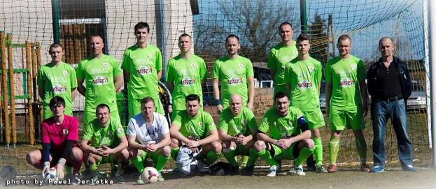 Eine Gruppe von Männern in grünen T-Shirts steht und hockt auf einem Fußballfeld mit einem Ball vor ihnen, ein Torpfosten rechts und ein Hintergrund aus Bäumen, Gebäuden, Masten, Fahrzeugen und Himmel, mit Text in der linken unteren Ecke, der die Zahlen von 1 bis 72 auflistet.