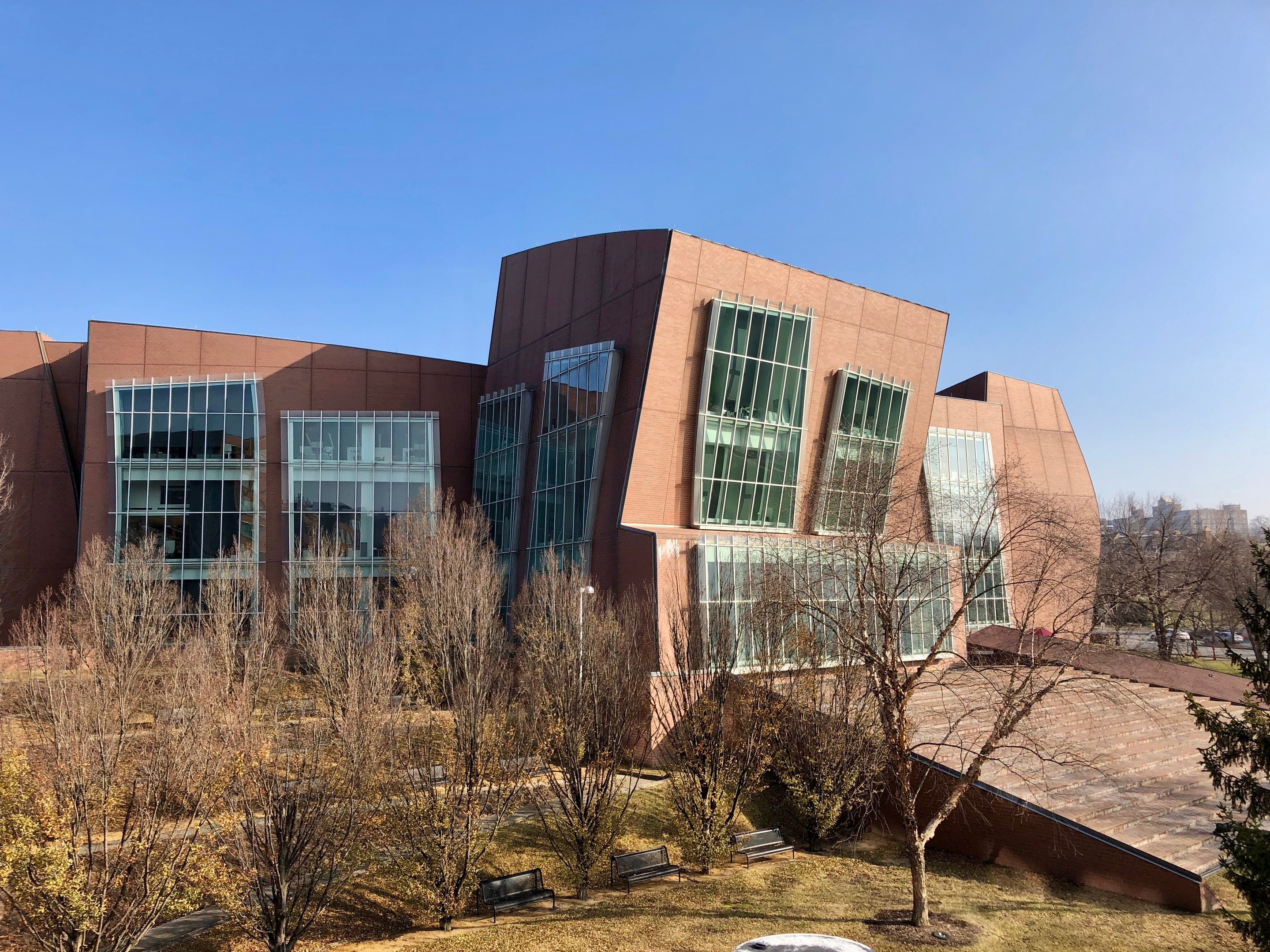 Großes Gebäude mit zahlreichen Fenstern, umgeben von Bäumen, Bänken, Gras und einem Laternenmast, mit anderen Gebäuden und einem klaren blauen Himmel im Hintergrund, als der neue Campus der University of Illinois at Chicago identifiziert.