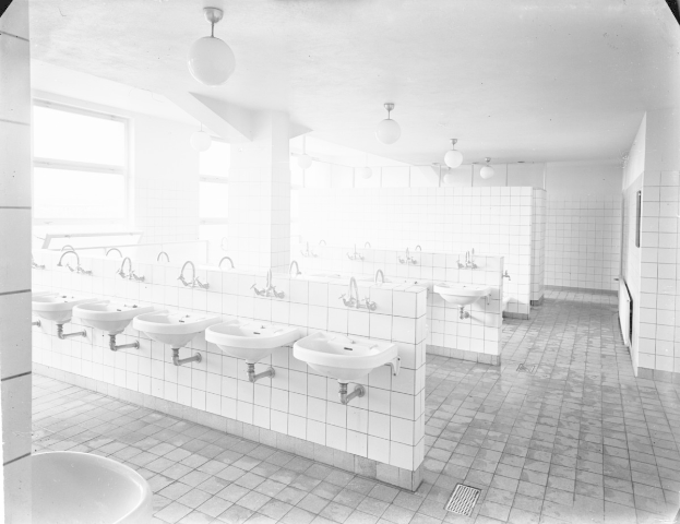 Schwarzes und weißes Foto einer öffentlichen Toilette mit Waschbecken, Wasserhähnen, Wänden, Beleuchtung und Fenstern.
