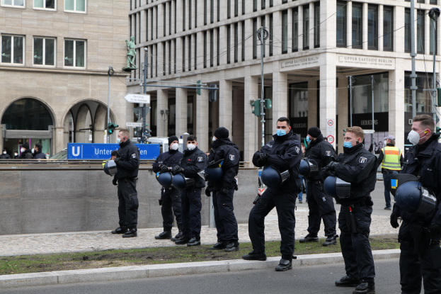 Eine Gruppe von Polizisten in schwarzen Uniformen und Masken steht vor einem glasfensterigen Gebäude mit Säulen in Berlin, Deutschland, einige halten Helme, mit Laternen, Verkehrsampeln, Texttafeln, einer Statue, anderen Menschen und grasbewachsenem Boden im Hintergrund.