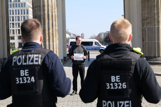 Drei Polizeibeamte stehen vor einem Gebäude, ein Mann in der Mitte hält ein Papier, flankiert von Säulen; Fahrzeuge, Menschen, Bäume und Gebäude sind im Hintergrund unter einem Himmel zu sehen, was auf eine nachrichtenbezogene Szene hinweist.