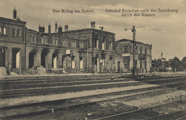 Schwarzes und weißes Foto des Bahnhofs Korschen in Ostern, Deutschland, mit Bahnschienen, Bahnhofsgebäuden, Menschen, Bäumen und einem Himmel als Hintergrund.