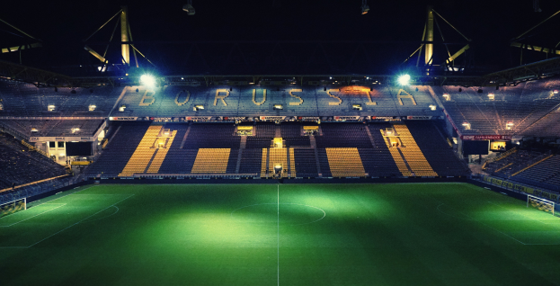 Ein Fuballfeld nachts, erleuchtet von hellen Stadionlichtern, mit einigen Menschen auf den R├Ąngen und saftig gr├╝nem Rasen.
