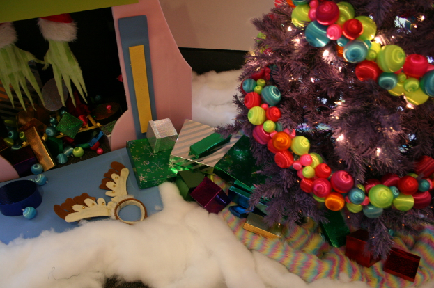Eine Indoor-Szene mit einer Wand im Hintergrund, einem Baumwoll- und Wolltuch unten, einem geschmückten Weihnachtsbaum rechts, Geschenkboxen und Dekorationsgegenständen in der Mitte und einer Hütte mit Dekorationen und Baumwolle oben links.
