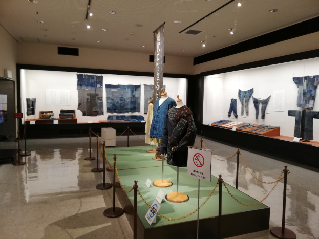 Ein Raum voller Schaufensterpuppen in traditioneller japanischer Kleidung, Vitrinen mit verschiedenen Kleidungsstücken, Informationsschilder und Deckenleuchten.
