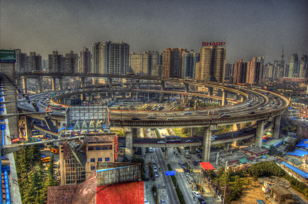 Stadtpanorama mit einer zentralen Ringstraße mit Fahrzeugen, einer unteren Straße mit Autos und Fahrrädern, zahlreichen Hochhäusern im Hintergrund, einem Himmel oben, Bäumen links unten und kleinen Häusern, die über die Stadt verteilt sind.