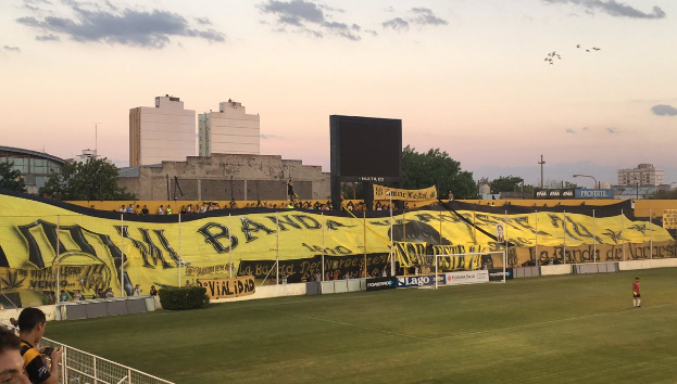 Ein Fußballfeld mit einer Gruppe von Menschen darauf, umgeben von einem Metallzaun, einem Torpfosten, einerBanner mit der Aufschrift "Boca Juniors vs Real Sociedad", einigen Pflanzen, einem Anzeigebildschirm, einigen Pfählen, der Rinde eines Baums, einigen Bäumen, einigen Gebäuden und einem bewölkten Himmel.