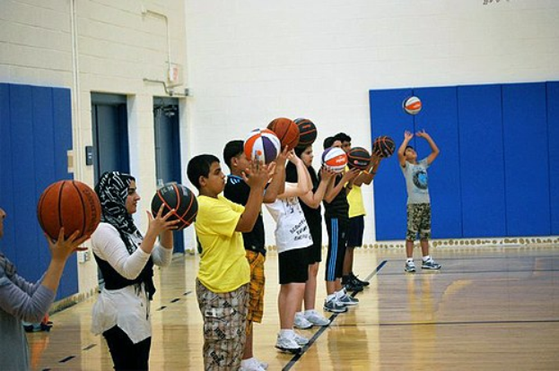 Gruppe junger Menschen mit Basketballs auf einem Basketballfeld bei einem Camp, mit Türen und einer Wand im Hintergrund.