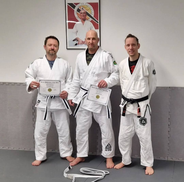 Drei Männer in weißen Judo-Unterhemden stehen nebeneinander in einem Fitnessstudio, jeder hält eine Urkunde in der Hand, mit einem Wandrahmen im Hintergrund, der darauf hindeutet, dass sie den ersten Platz im Judo gewonnen haben.
