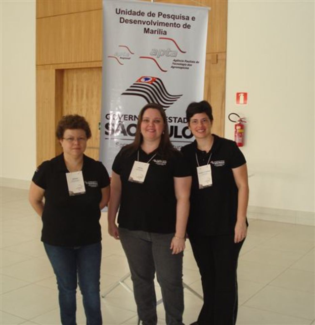Drei Frauen in schwarzen T-Shirts stehen zusammen, zwei lächeln, mit einem Banner und einem Feuerlöscher an der Wand dahinter.