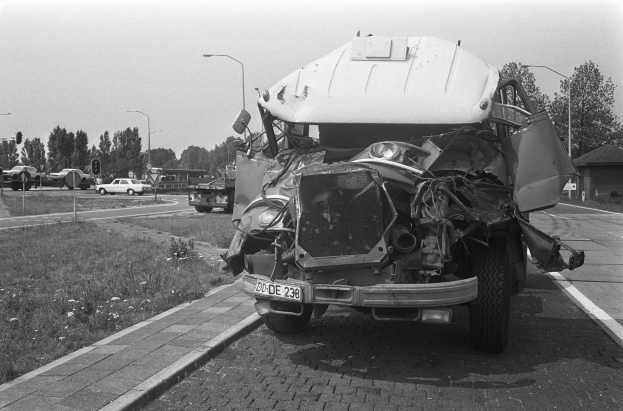 Schwarz-weiß-Foto eines schwer beschädigten Autos am Straßenrand mit eingedellter Motorhaube und zersplitterten Fenstern, umgeben von anderen Fahrzeugen, Laternenpfählen, Bäumen und einem Haus.