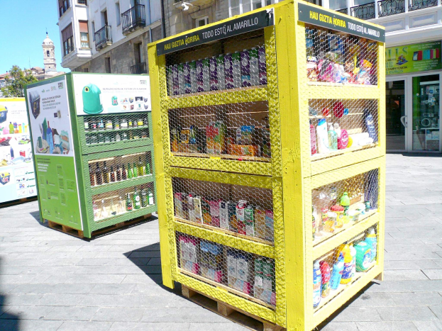 Ein gelber Verkaufsautomat auf einer Stadtstraße, gefüllt mit Gegenständen, umgeben von Gebäuden, Bäumen und einem klaren blauen Himmel.